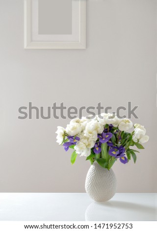 Beautiful bouquet of white peonies and purple irises in the living room on a glass table on a background of gray wall