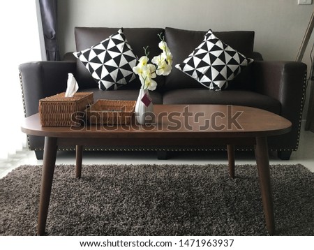 Square pillow on a fancy, black sofa and a basic, wooden coffee table on a brown brahma in a white living room interior. Real photo.