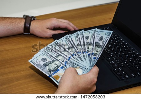dollars in male hands and a laptop on the background of a wooden, office table.
