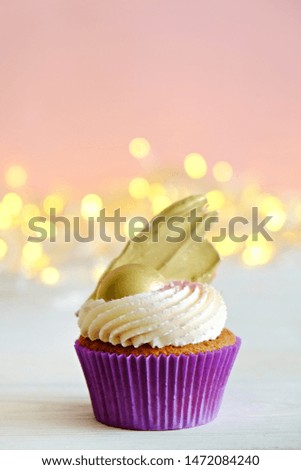 Sweet organic cupcake in purple wrap with cream cheese frosting swirl and golden chocolate decoration on white wooden textured table. Close up, copy space, background.
