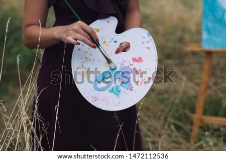 woman with paints palette and brush in hands standing on meadow painting outdoors