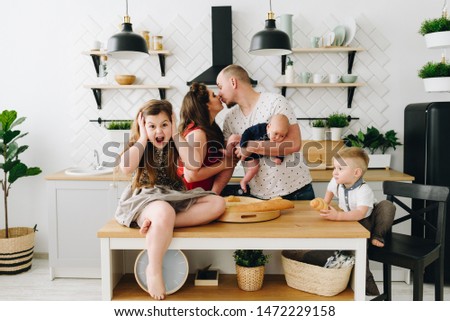 A little girl sitting on the kitchen table and screaming while her parents are kissing behind and brother is holding a bun 