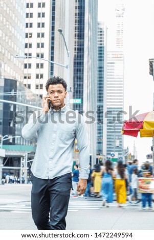 Young Mix Race American Man talking on cell phone, traveling in New York City, wearing long sleeve gray shirt, walking on street in Midtown of Manhattan. Modern high buildings and people on background