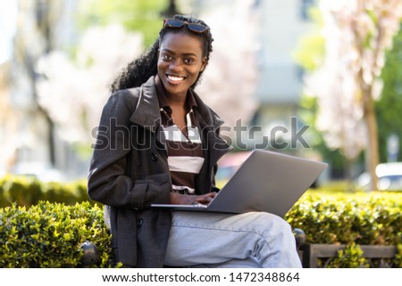Beautiful black business woman sitting on bench in the financial city, using a laptop computer, thoughtful, sunny outdoors.