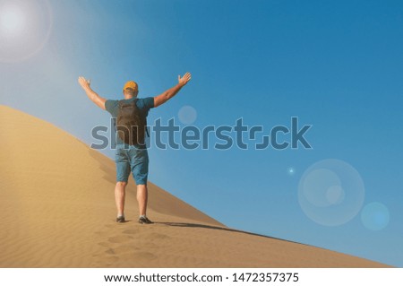 A man with backpack on the sand dune happy to be there. Male rise his hands in desert landscape