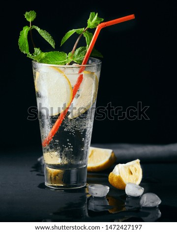 Homemade lemonade, detox water or alcoholic drink. A glass with lemon and mint and ice on dark black background with red straw. Copy space, place for text.