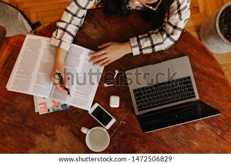Top view of girl reading book and taking notes in it. Woman sitting at table with laptop