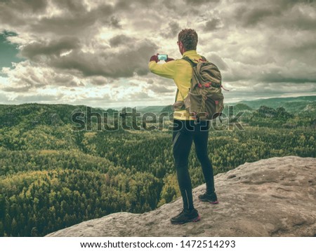 Man tourist hiking mountain trail, takes picture by phone. Hiker walking on rocky hill, wearing backpack and sunglasses, using trekking sticks