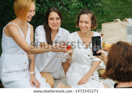 Three young women in white clothes during picnic. They are holding glasses of rose wine, making a toast, while forth is taking a picture of them on her phone.