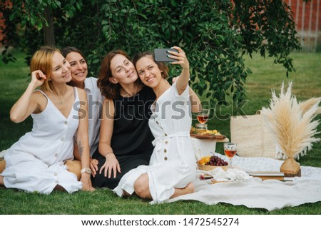 Four young women are taking selfie during picnic on a lawn under tree. Three are dressed in white and one in black. They are sitting next to wine and variety of appetizers set on a white tablecloth.