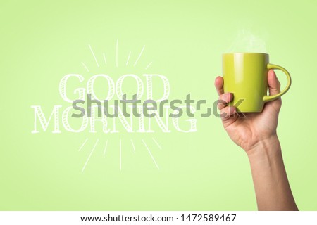 Male hand holding a green cup with hot coffee or tea on a light green background. Added text Good morning. Breakfast concept with hot coffee or tea
