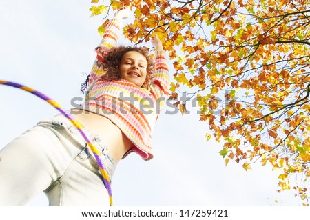 Low view of an active teenager girl playing hoola hoop in a park with orange and brown leaves on the trees, being joyful, smiling and playing during a sunny autumn day.
