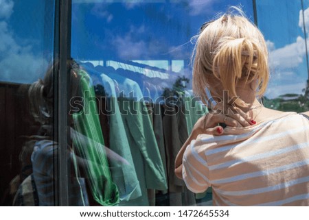 A young blonde girl with a hairpin on her head in a striped T-shirt is looking at shop windows with clothes on a sunny day, a photo from the back
