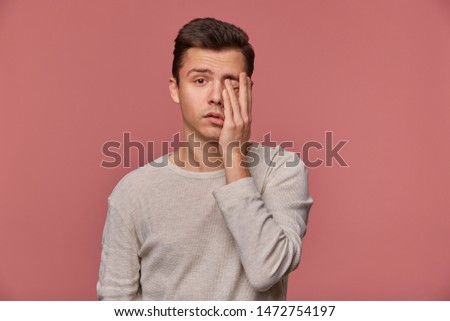Portrait of young attractive man in white long sleeve, looks at the camera and touches face, stands over pink background, looks tired and sad.