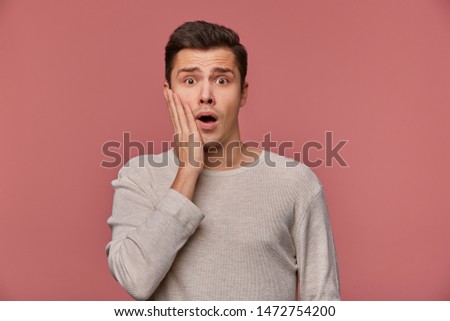 Young handsome dazed guy wears in checkered shirt, looks at the camera with wide open mouth in surprised expression, touches cheek, isolated over pink background.