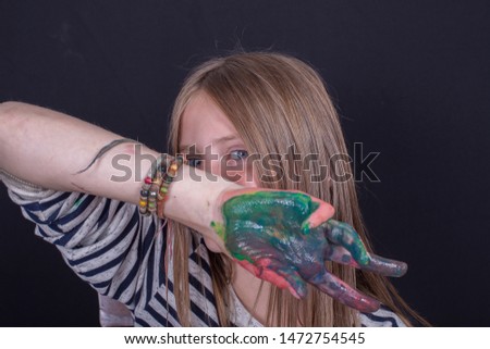Beautiful blond young girl with freckles and hands painted in colorful paints indoors on black background, closeup portrait