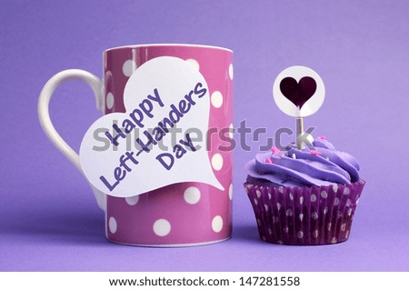Happy Left Handers Day message written on white heart shape tag on pink polka dot mug with purple cupcake on purple background, for International Left-Handers Day celebrated on August 13. Royalty-Free Stock Photo #147281558