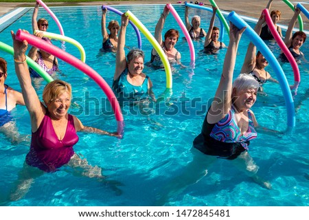 Action portrait of senior aqua gym class. Elderly ladies exercising together with foam noodles in outdoor swimming pool. Royalty-Free Stock Photo #1472845481