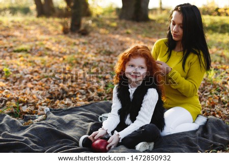 Autumn vibes, family portrait. Charming mom and her red hair daughter have fun sitting on the fallen leaves in the park full of evening sun