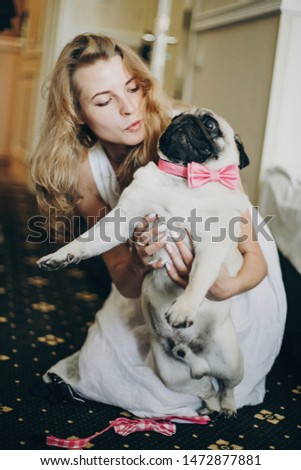 Cute bride hugging funny pug dog with pink bow tie in morning  before wedding ceremony in hotel room.Girl with her dog. Pets at wedding day.