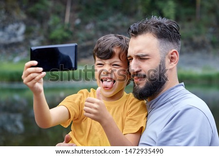 Father and son take selfies in nature, using a mobile phone camera, the boy is grimacing and showing his tongue.