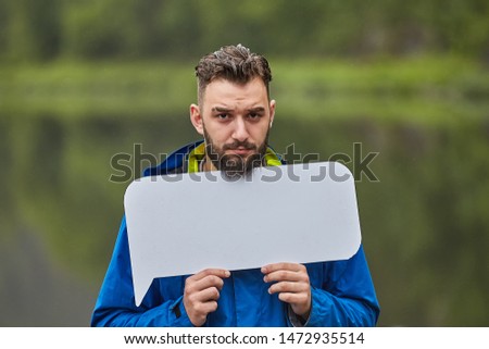 Upset young man holds a blank poster with a mockup for your advertisement on the background of nature.