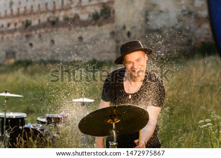 A musician in a black hat plays the drums emotionally, with water droplets on the tools. Around it the nature and ruins of the castle
