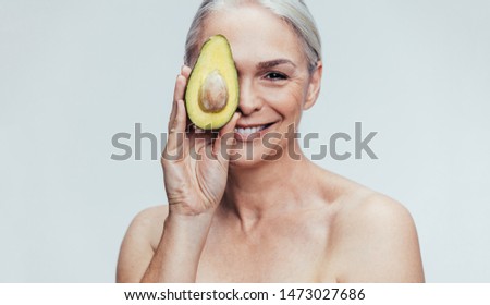 Happy senior woman holding half of fresh ripe green avocado in front of her eye against grey background. Female showing avocado to the camera.