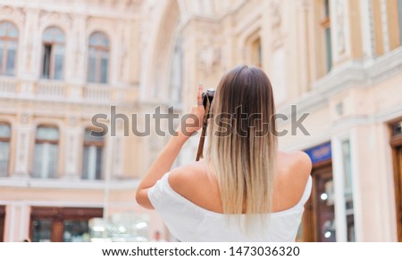 Blonde pretty photographer woman with retro camera in her hands while taking pictures at urban old architecture. Discover new places. Back view