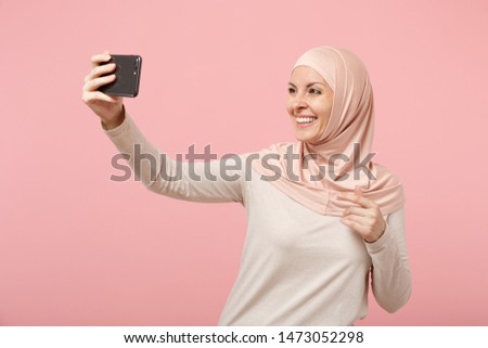 Smiling young arabian muslim woman in hijab light clothes posing isolated on pink background in studio. People religious Islam lifestyle concept. Mock up copy space. Doing selfie shot on mobile phone