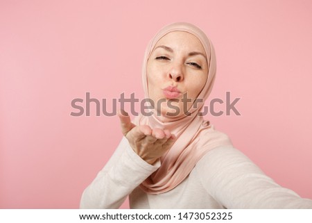 Close up arabian muslim woman in hijab light clothes posing isolated on pink background. People religious lifestyle concept. Mock up copy space. Doing selfie shot on mobile phone, sending air kiss