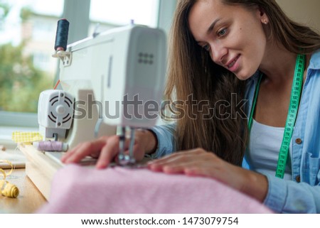 Happy smiling seamstress with electric sewing machine and different sewing accessories during tailoring process at workplace 