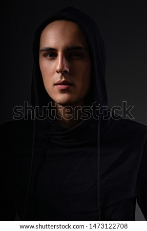 Mysterious serious man in black hoodie with hood on the head on dark background. Dangerous criminal person in dark shadow