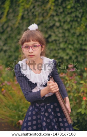 Schoolgirl with a big brown notebook on a background of greenery. Little girl 9 years old in school clothes. A girl with blond hair and a white hairpin. Blue and white clothes. Back to school