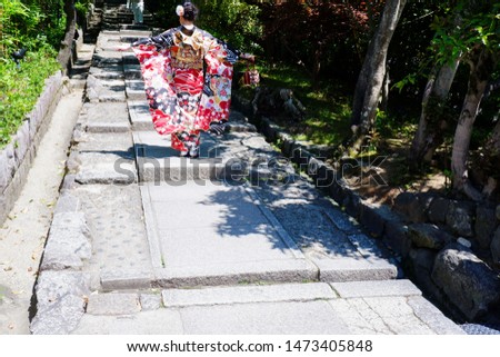 JAPANESE YOUNG WOMAN IN COLORFUL KIMONO SPREADING HER HANDS ON SUNNY STONE PARK STAIRS