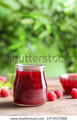 Glass jar of sweet jam with ripe raspberries on wooden table against blurred background