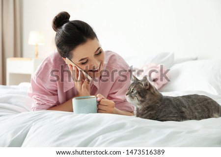 Young woman with cup of coffee talking on phone while lying near cute cat in bedroom. Pet and owner
