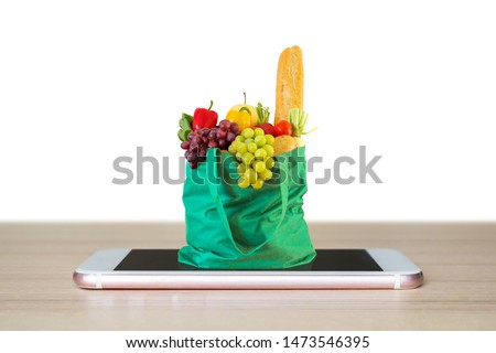 Fresh food and vegetables in green shopping bag on mobile smartphone on wood table top isolated on white background grocery online concept