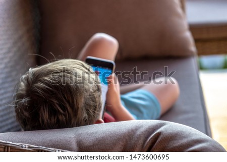 Back view image of unrecognizable little boy playing games on smart phone via social networks, sitting in home interior. 
Close up,young boy watching video, during vacation with his family. 