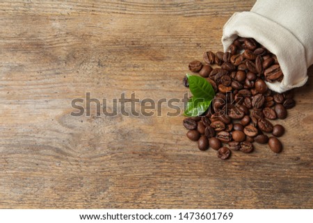 Bag of coffee beans and fresh green leaves on wooden table, top view with space for text
