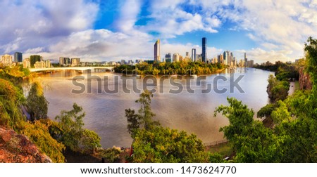 Green fresh Cliffs park on shores of Brisbane river across city CBD high-rise towers in wide panorama on sunny morning under blue sky.