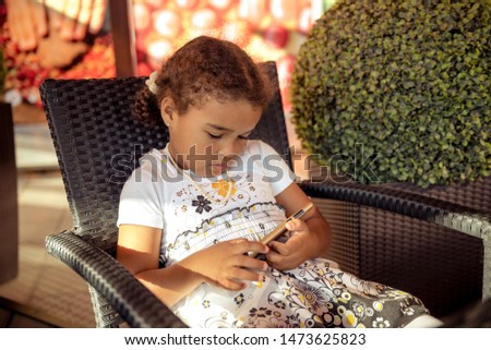 Beautiful little girl playing game or watching video on smartphone mobile. Girl watching cartoons or browsing internet, copy space. Side view portrait of girl using smartphone while sitting.