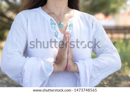 Woman in namaste gesture close up, practicing yoga and meditation outdoors. White clothing with boho chic jewelry. Beautiful soft summer light with nature background.