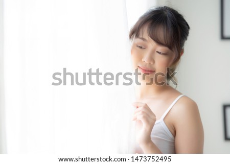 Portrait of beautiful young asian woman standing look at the window and smile while wake up with sunlight at morning, girl happy with fresh and cheerful, lifestyle and relax for wellness concept.