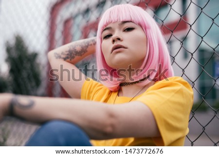 Stylish Asian teen influencer in pink wig looking at camera while sitting near net fence on blurred background of city street Royalty-Free Stock Photo #1473772676