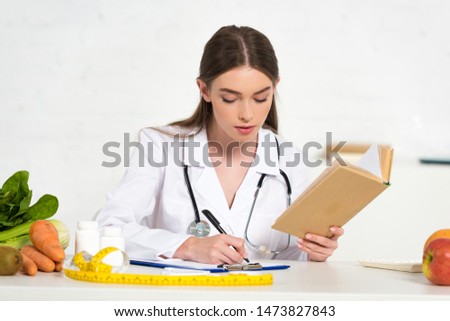 focused dietitian in white coat reading book and writing in clipboard at workplace