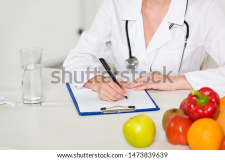 cropped view of dietitian in white coat writing in clipboard at workplace