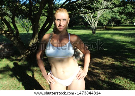 Blond Woman posing wearing a white bikini with her hair wet under the trees with sun in her face and trees and green grass in the background