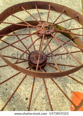 Two old rusty wagon wheels with metal rims and spokes abandoned on the ground in a state of decay. A square of orange paint in the bottom corner compliments the brown, beige and red colours.
