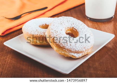 Donuts covered with icing sugar on a plate on a wooden table accompanied by a glass of milk and a fork on an orange napkin. Delicious breakfast or lunch. American food. Bakery and pastry products.